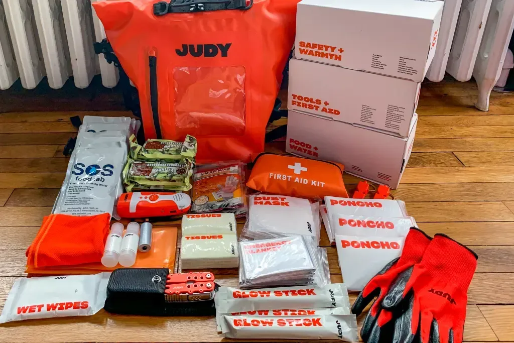 Emergency supply kits and food items arranged on a wooden floor, with orange bags and a white box.