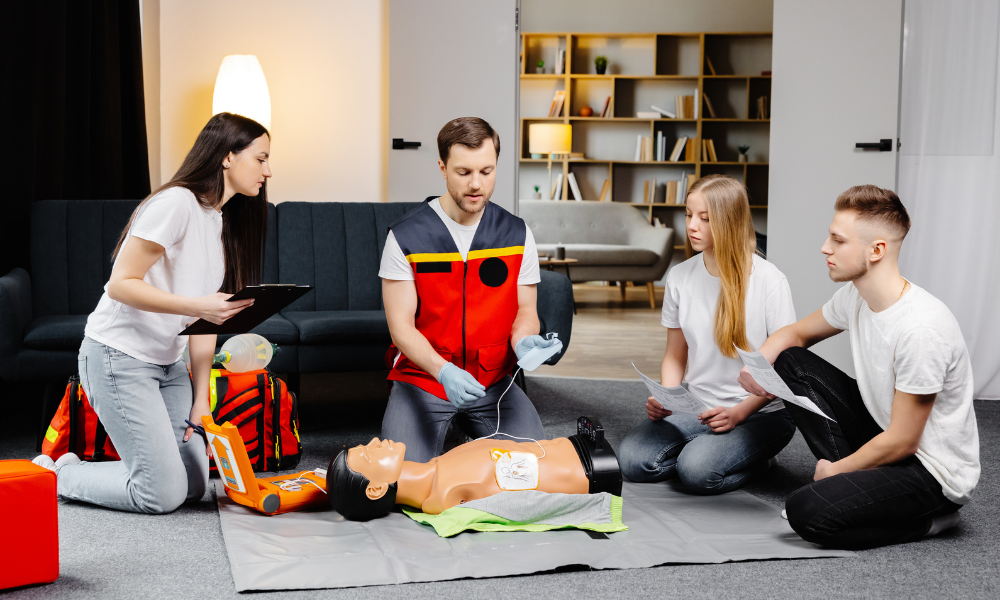 First aid training with a man practicing CPR on a mannequin while others watch and assist in a living room.