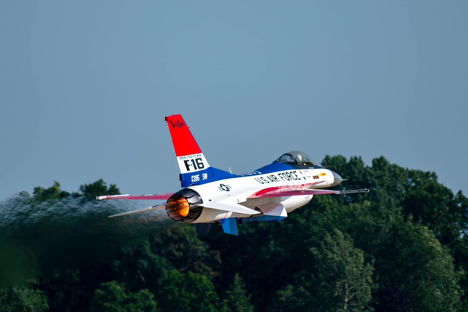 Red, white, and blue fighter jet flying low over trees against a clear blue sky