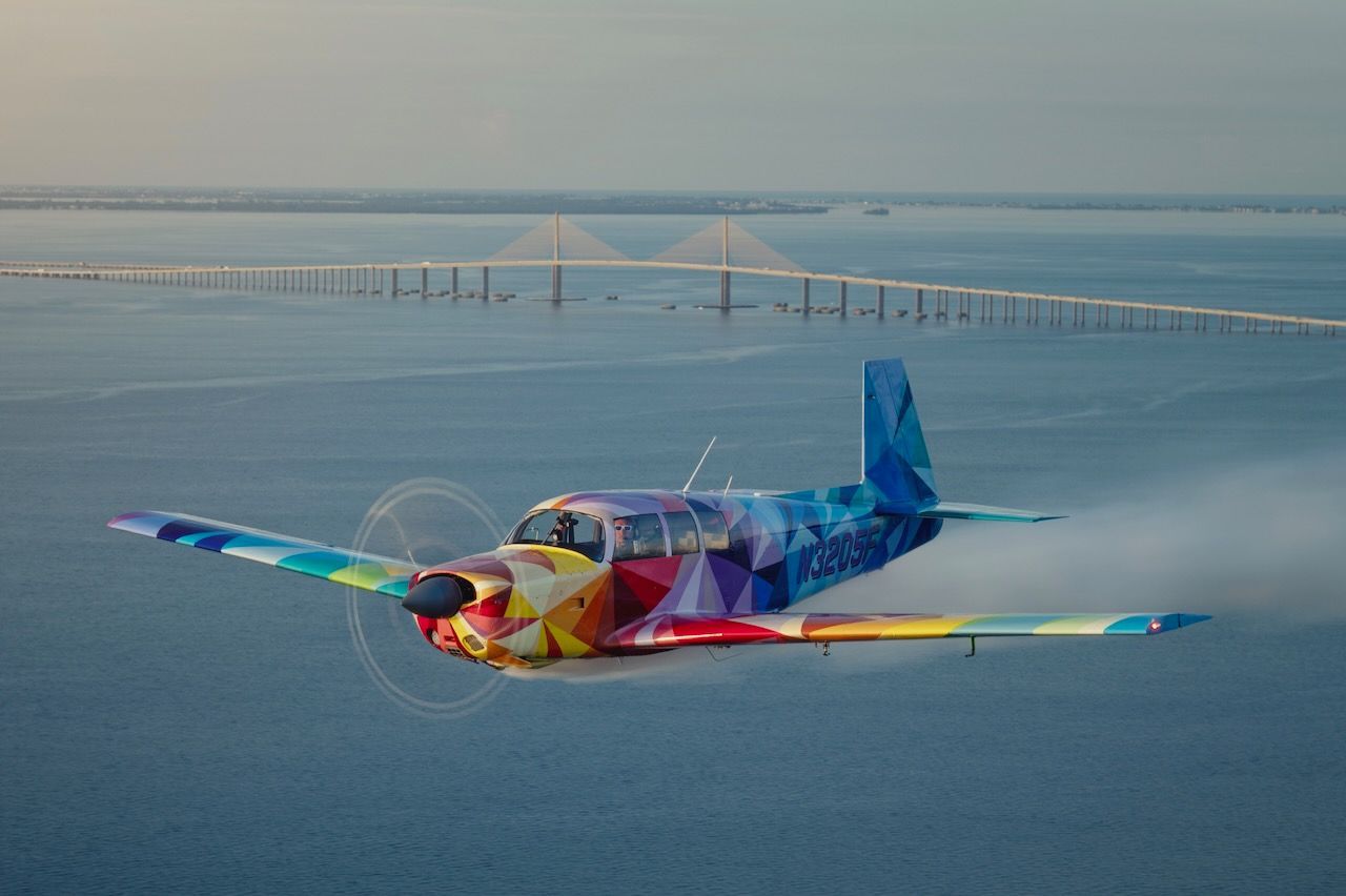 Colorful geometric airplane flying low over water with a long bridge in the background