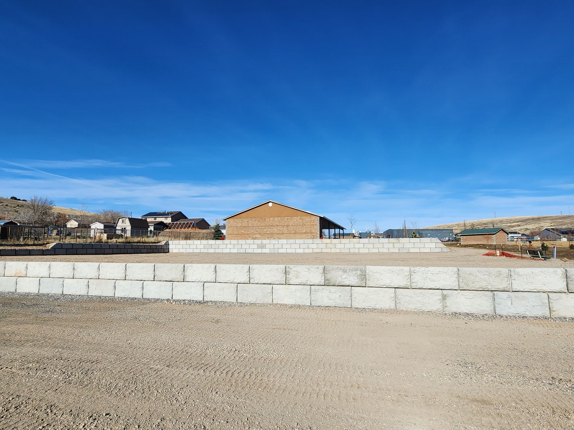 Stone wall and gravel lot with a tan building under a clear blue sky.
