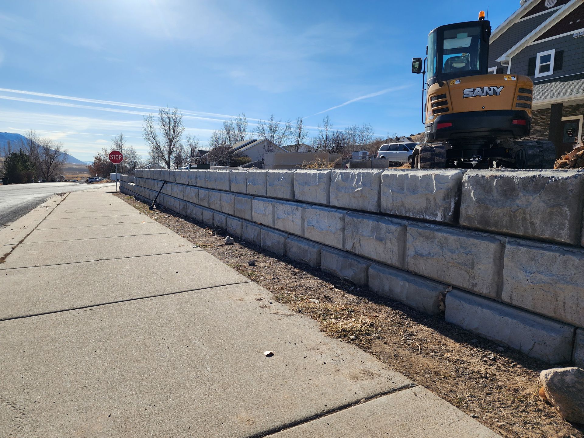 Sidewalk beside a stone retaining wall under construction with an excavator on top under a blue sky