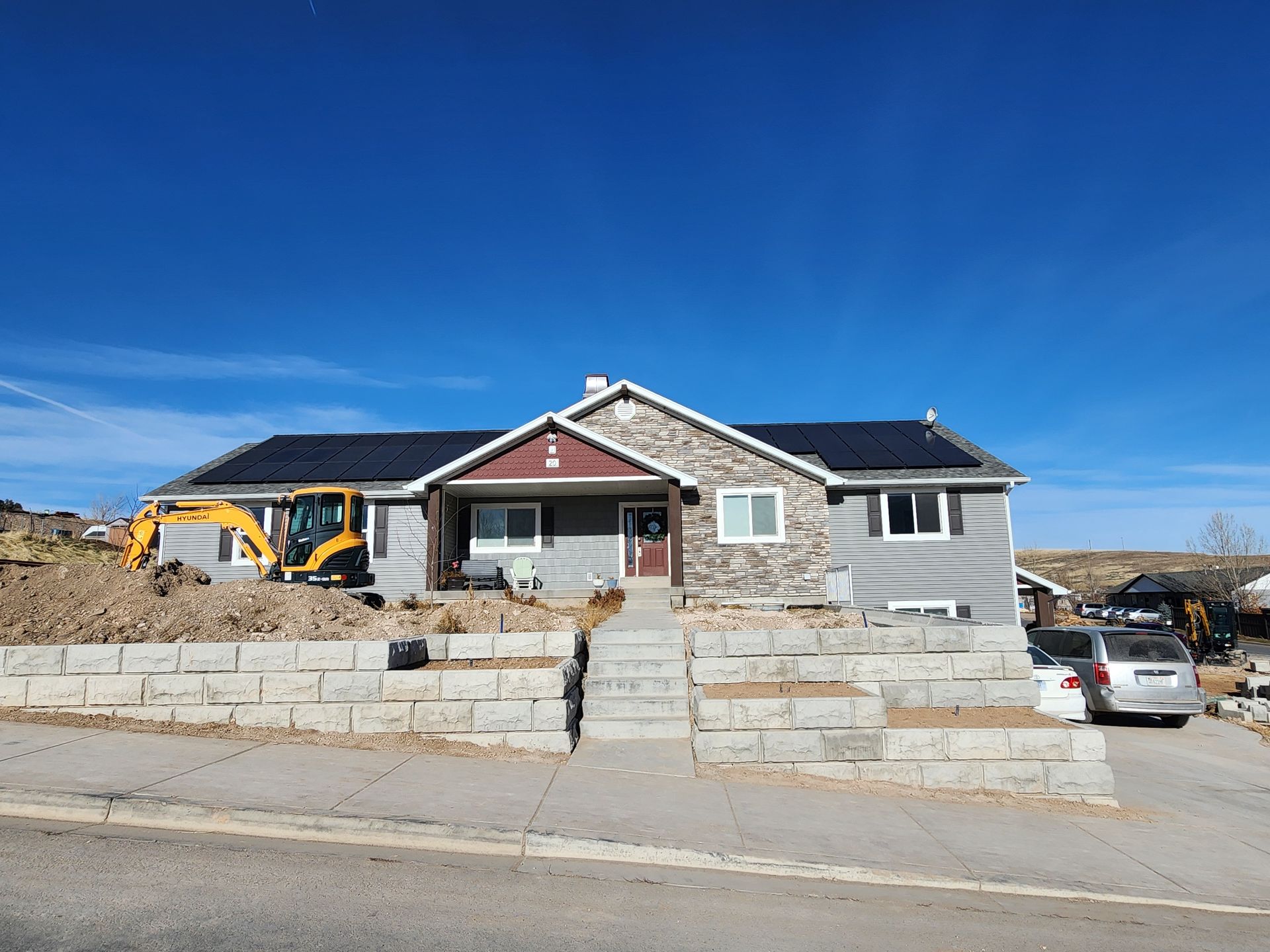 Single-story house under construction with a yellow excavator, stone retaining wall, and parked car on a clear day