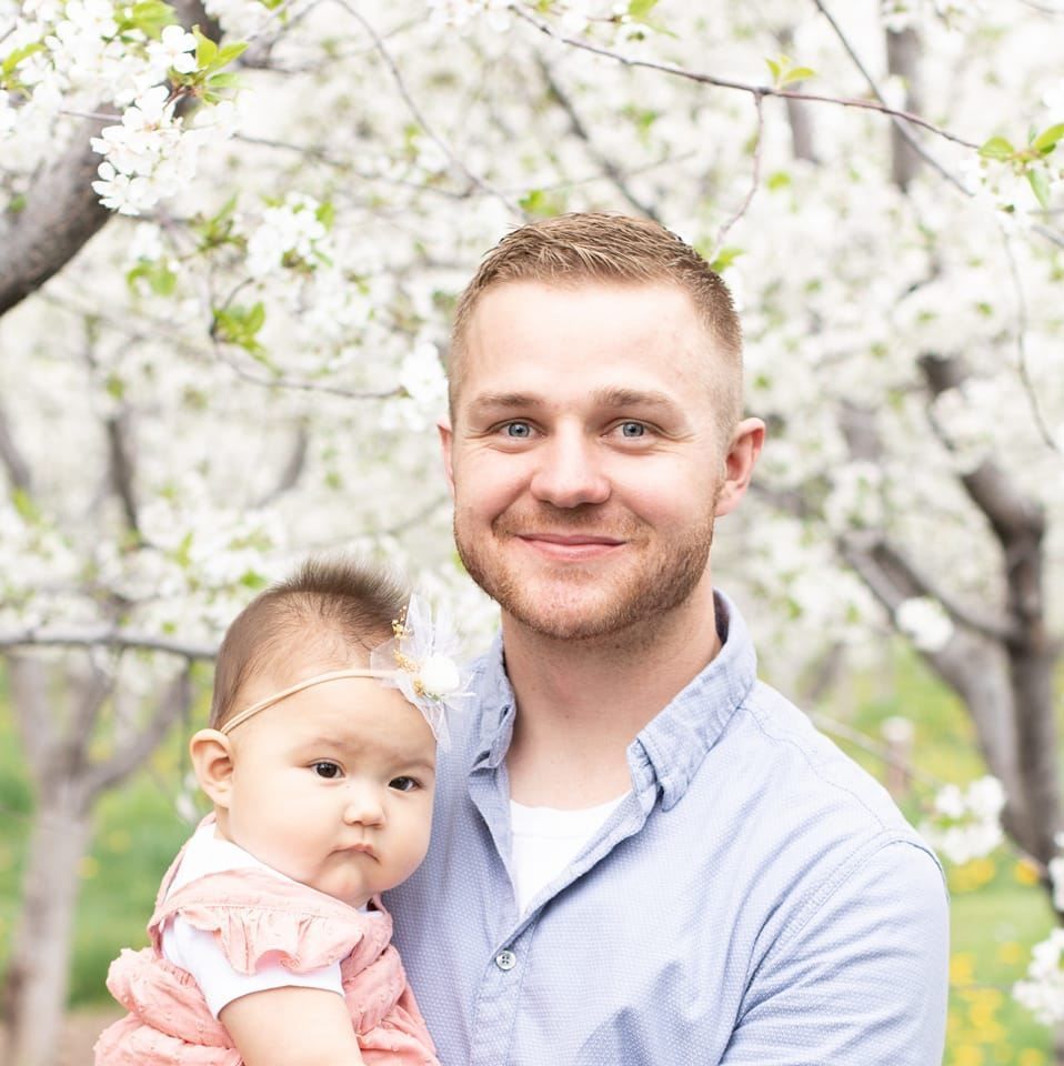 Man holding a baby in a blossoming orchard, both facing the camera and smiling.