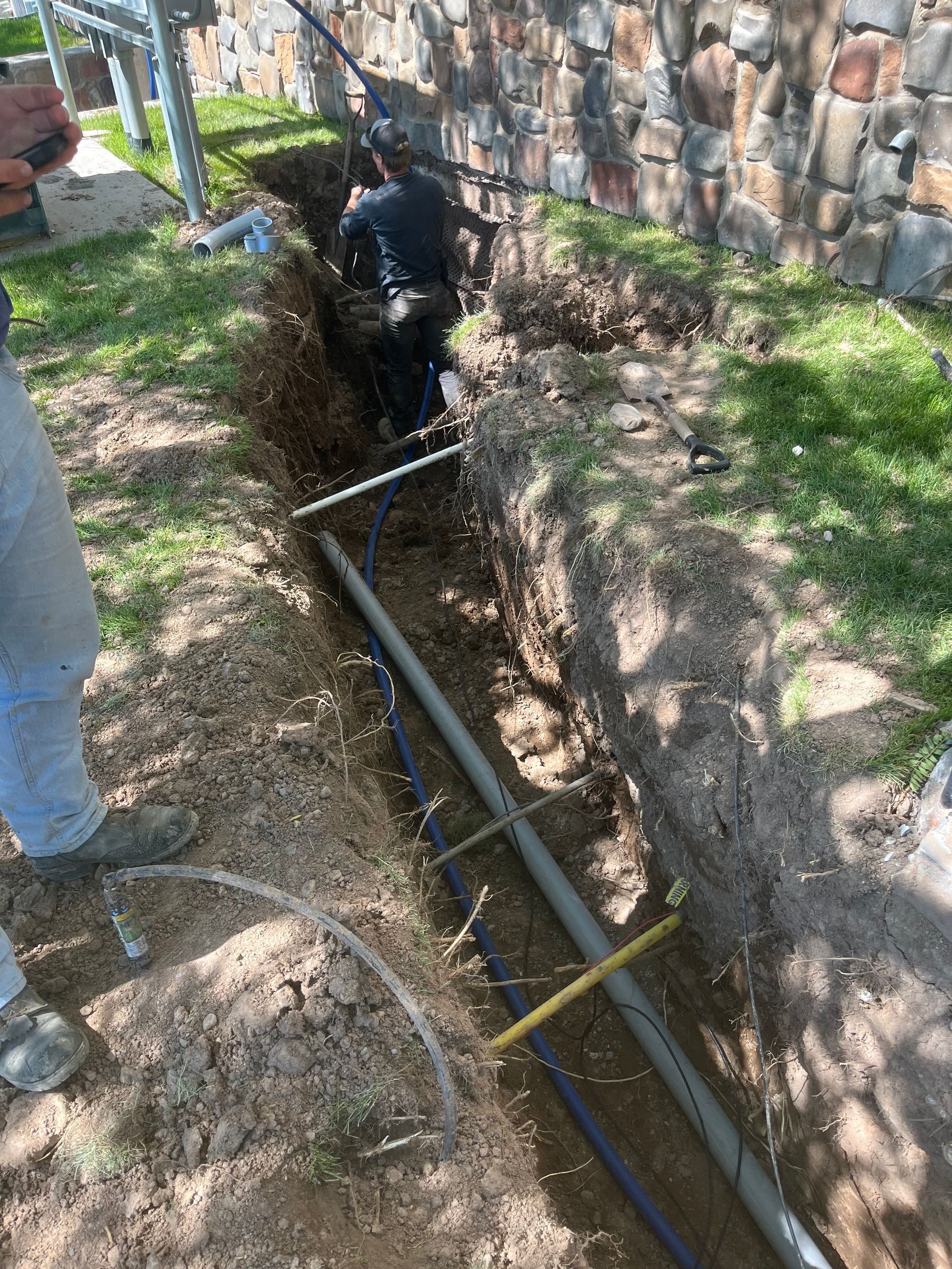 Worker digging a trench beside a stone wall, with pipes laid in the dirt and grass nearby