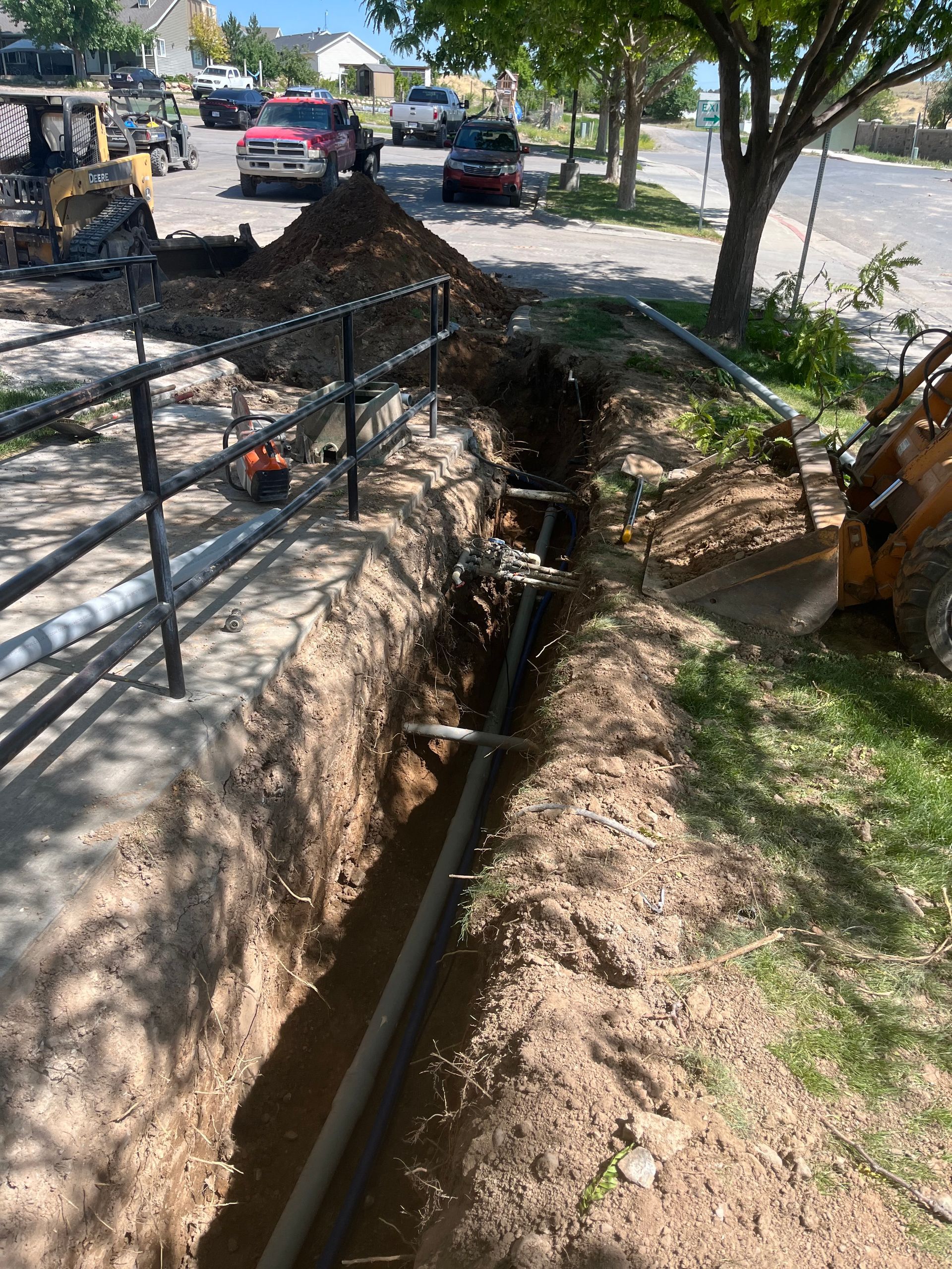 Open roadside drainage ditch beside a sidewalk, with cars and trees along the street.