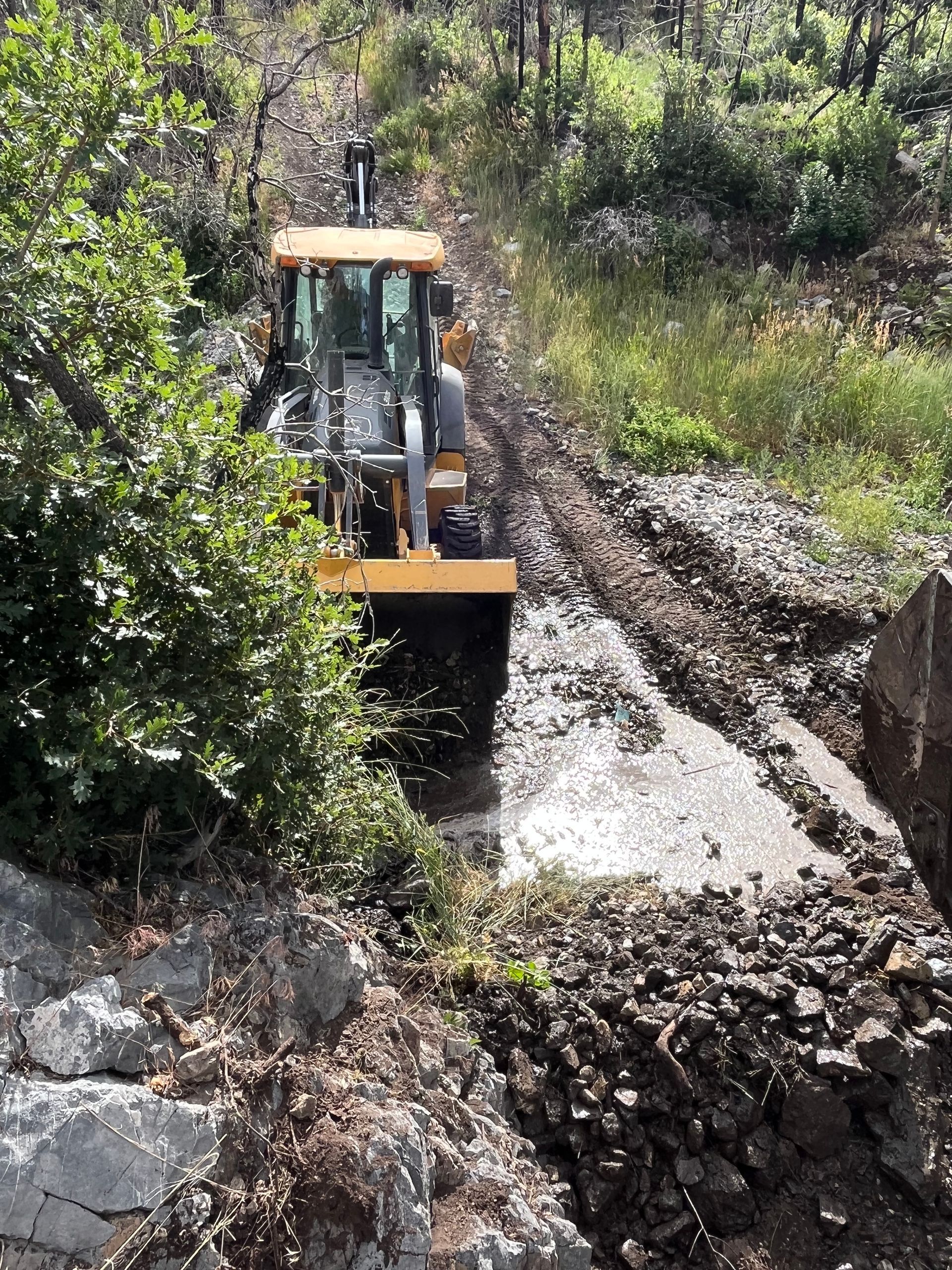 Backhoe clearing mud from a narrow dirt path in a wooded area