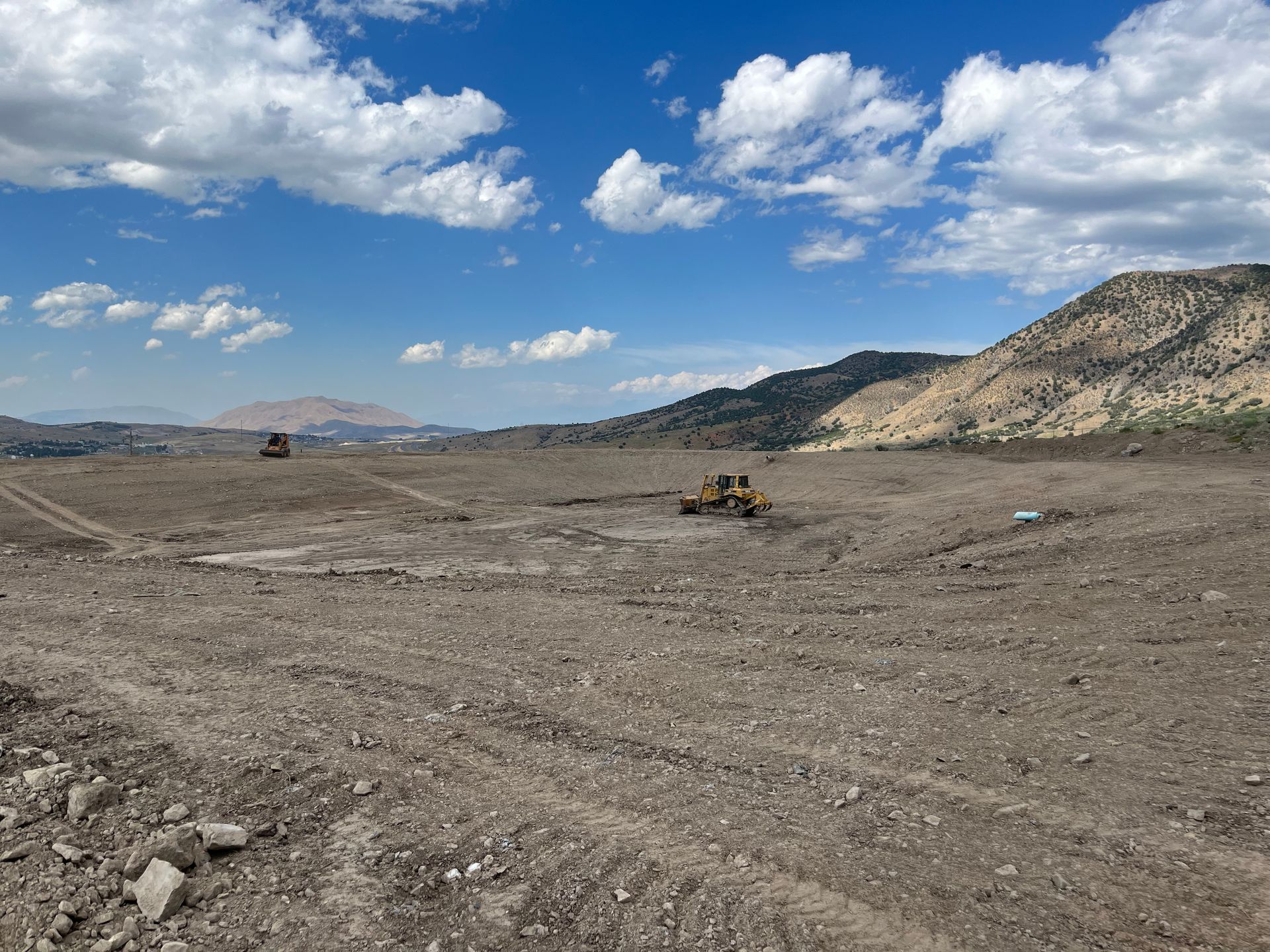 Wide barren construction site with bulldozers under a blue sky and rocky hills in the background