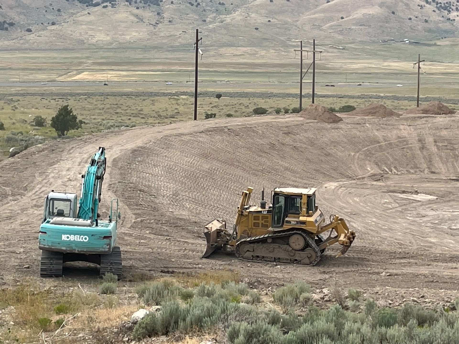 Excavator and bulldozer working on a dirt road in a barren rural landscape, with hills and power poles behind.