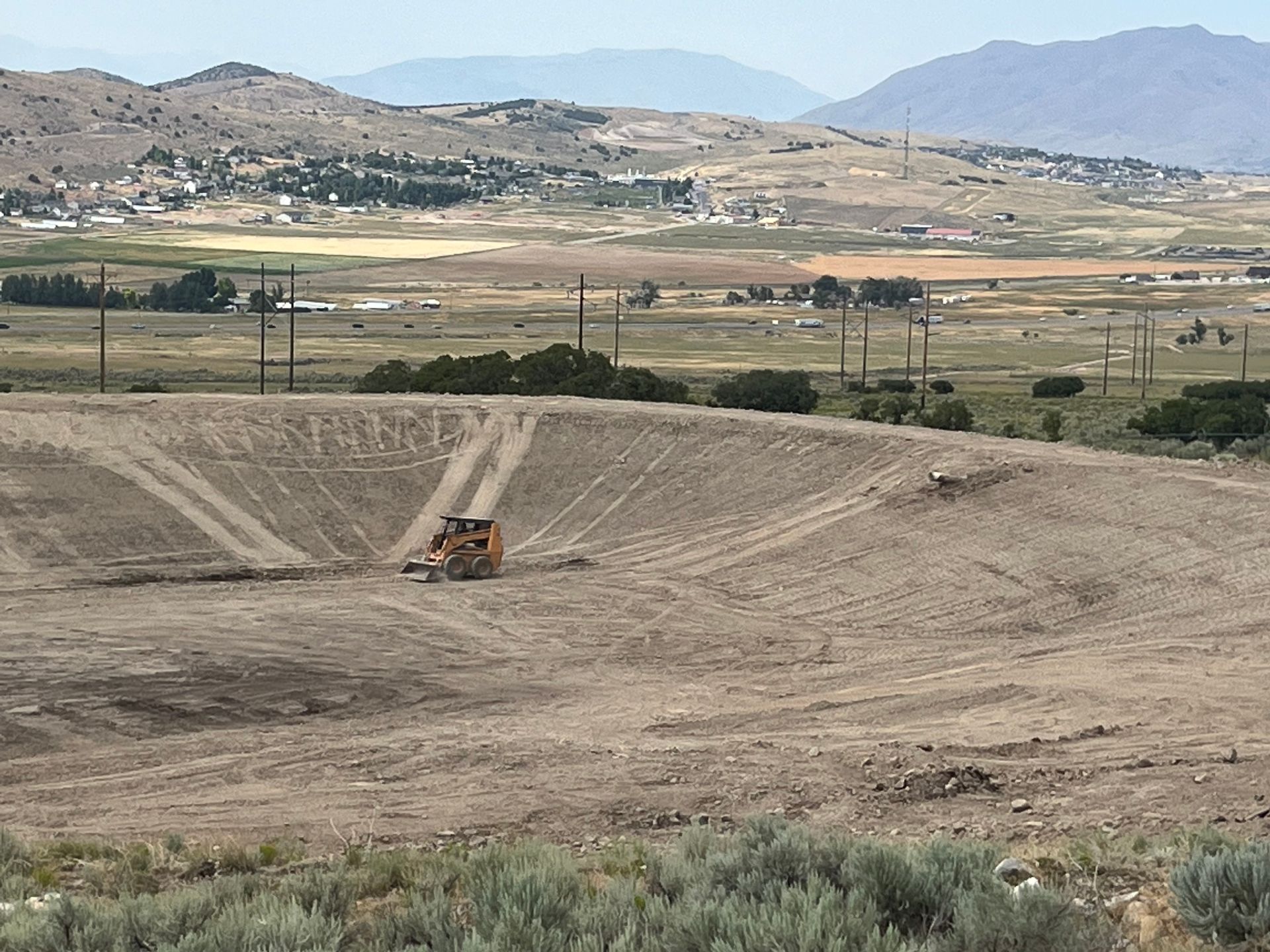 Wide dry dirt basin with a small bulldozer, fields, and distant mountains under a hazy sky