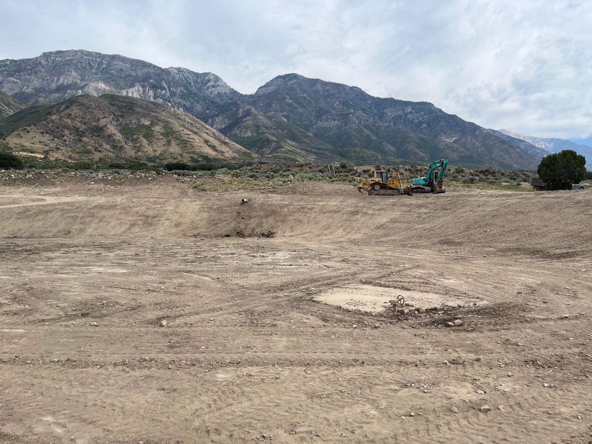 Empty dirt lot with an excavator near mountains under a cloudy sky