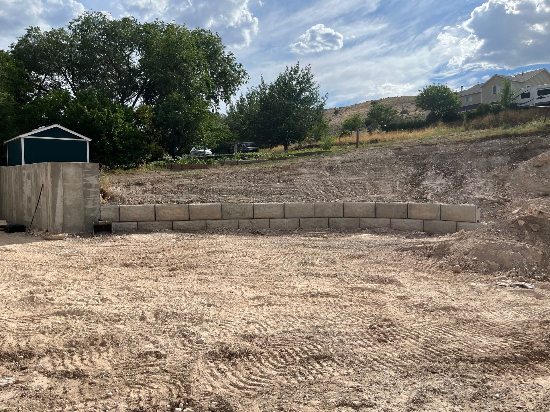 Bare dirt lot with a cinder block retaining wall, trees, and a small shed under a cloudy sky