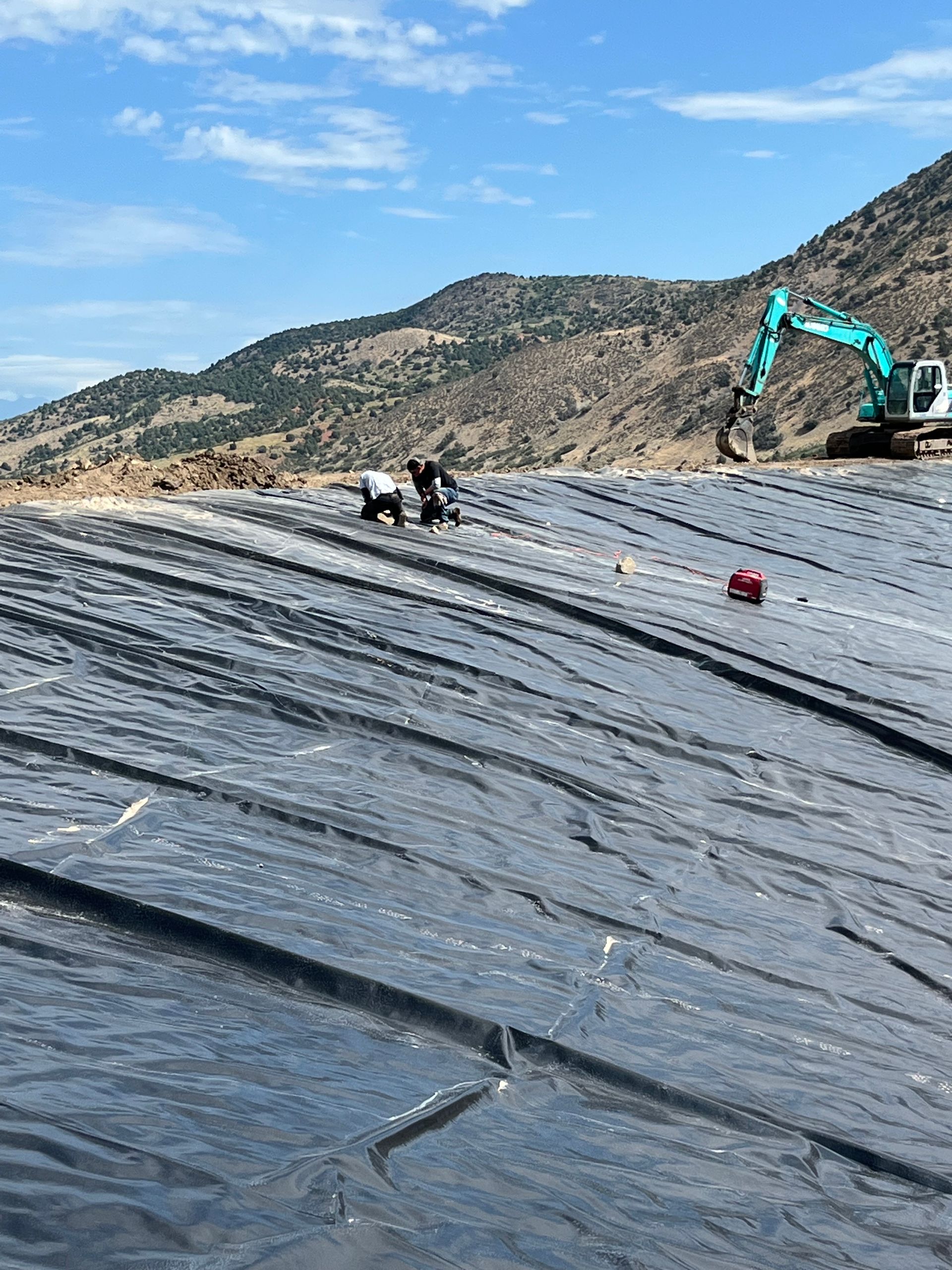 Black plastic-lined slope at a construction site with workers and an excavator under a blue sky