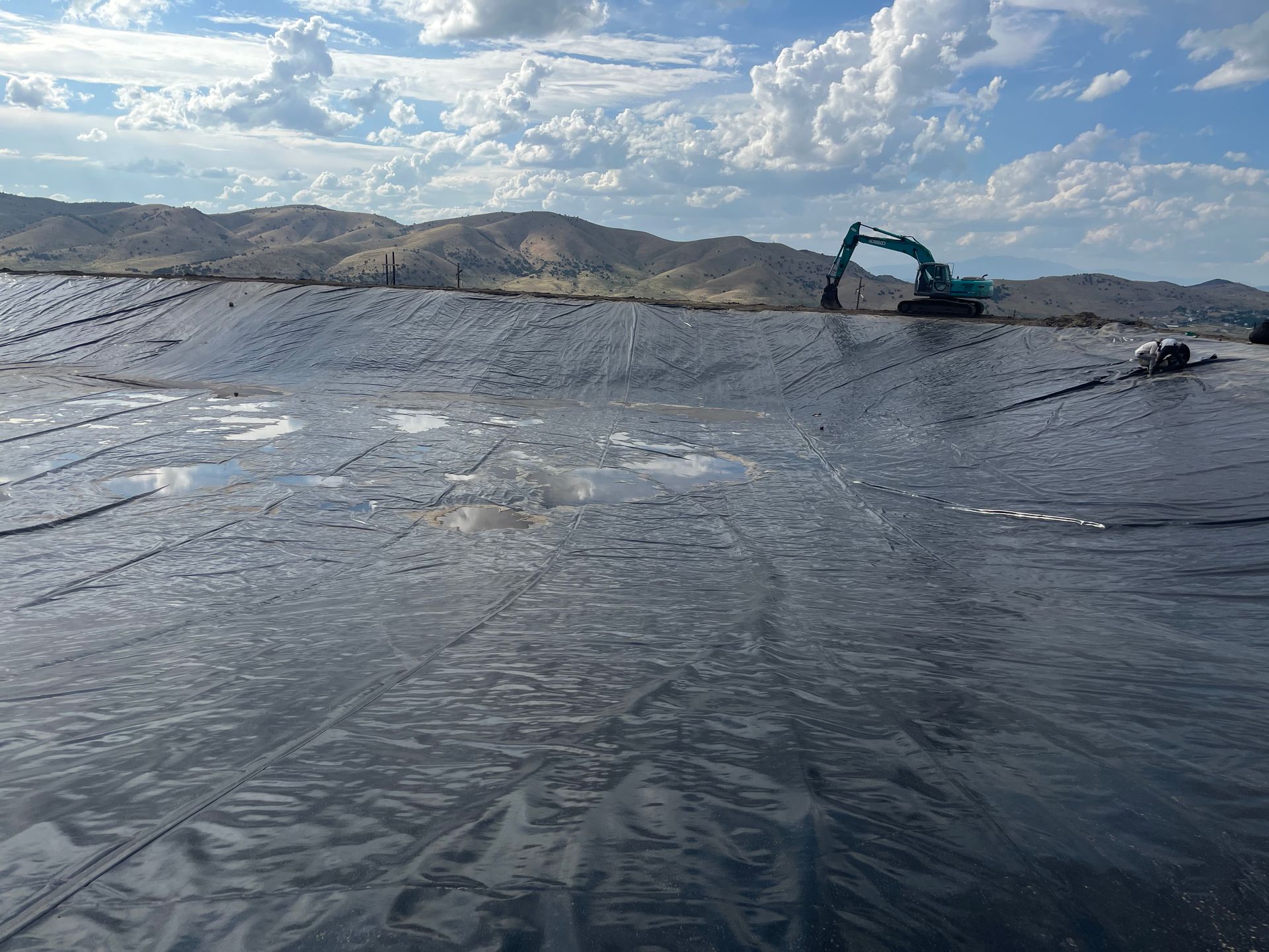 Excavator working on a large black plastic-lined pond under a cloudy sky, with hills in the background