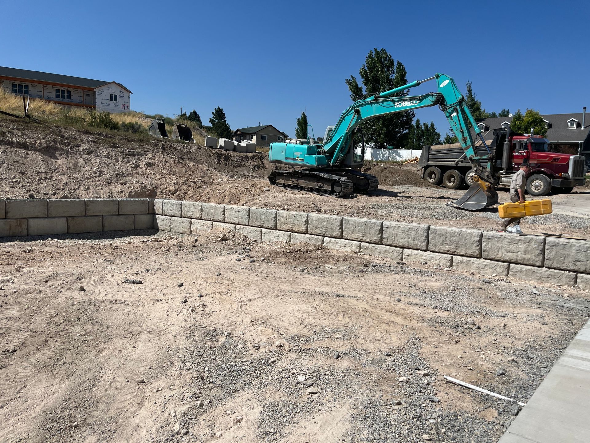 Excavator and dump truck at a construction site with stone retaining walls under a clear blue sky