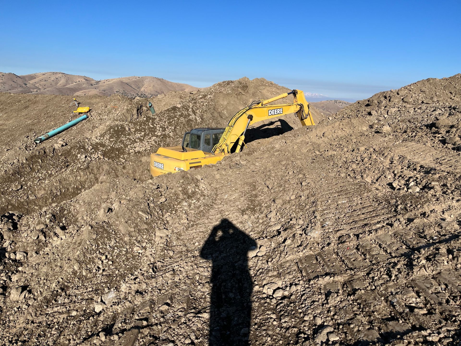 Yellow excavator on a rocky hillside with a person’s shadow in the foreground.