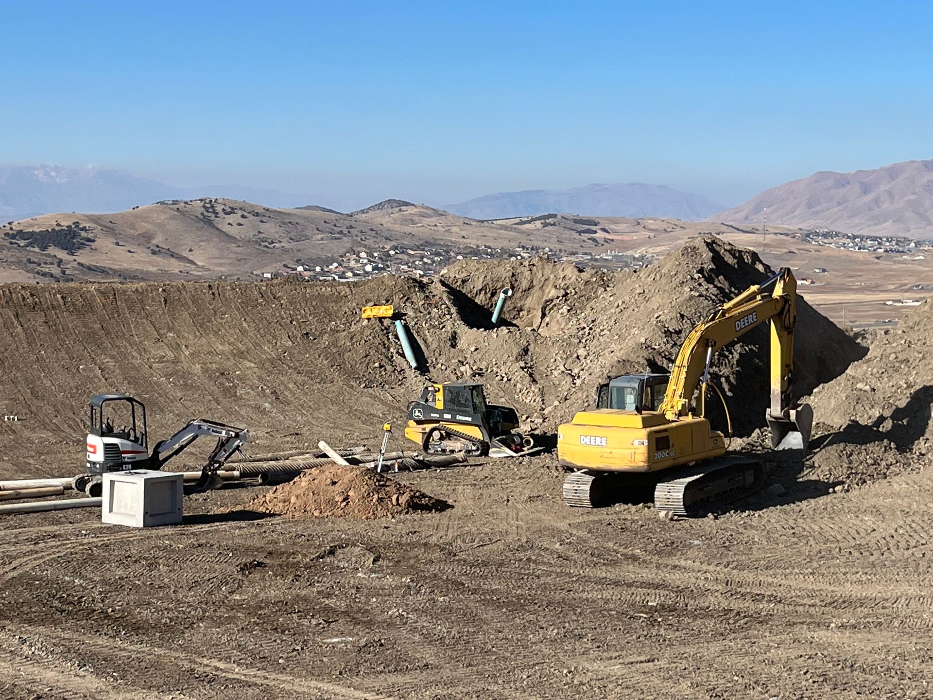 Two yellow excavators and a small loader at a dusty construction site with hills in the background