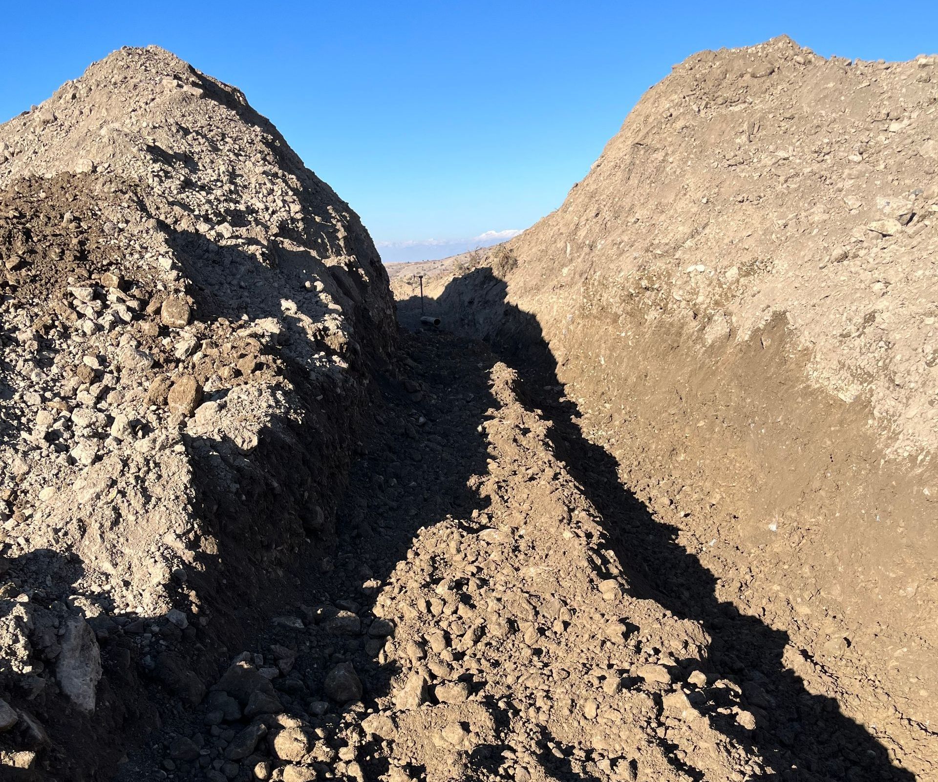 Large dirt piles forming a narrow trench under a clear blue sky.