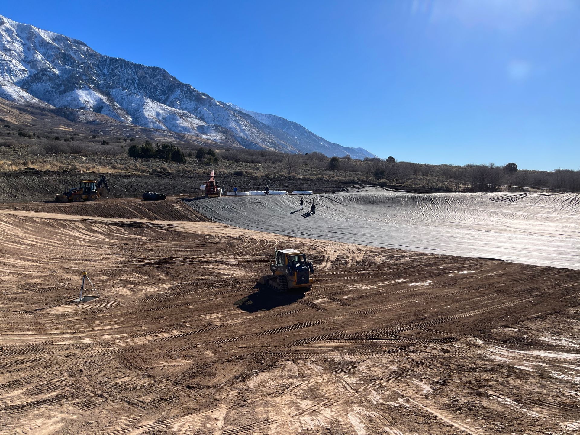 Excavator grading dirt beside a reservoir with snowy mountains under a blue sky