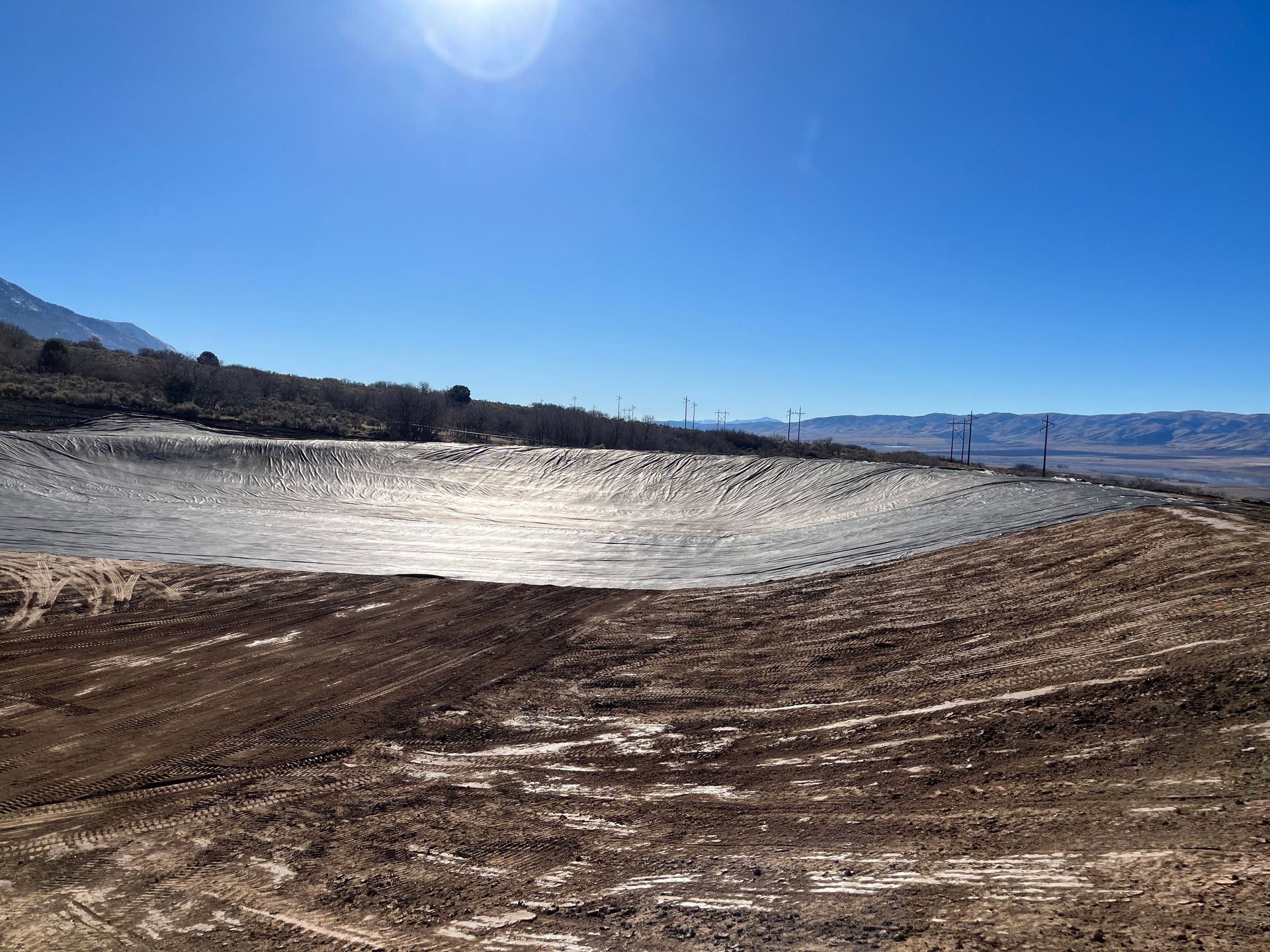 Bright blue sky over a dry, muddy lakeshore with distant mountains and bare trees
