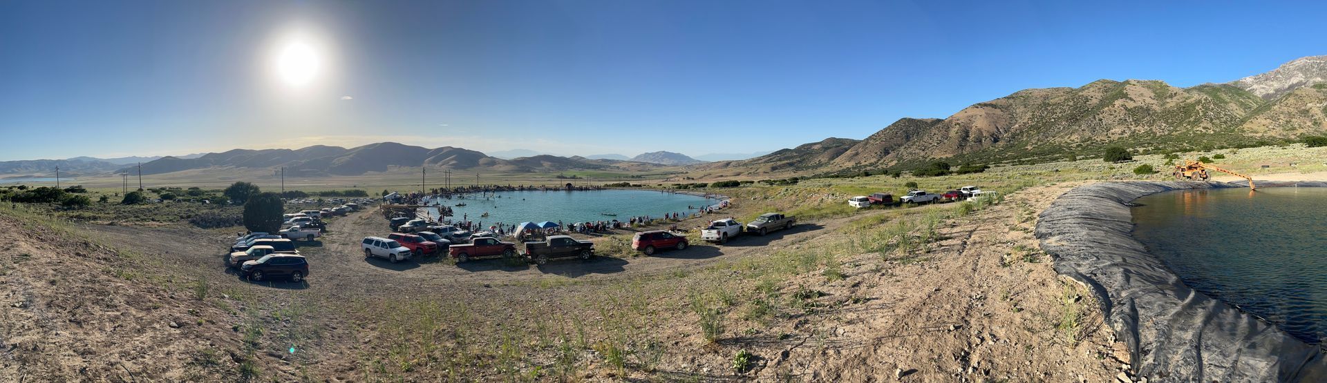 Cars and tents on a rocky lakeshore under a bright sun, with hills and blue water in the background