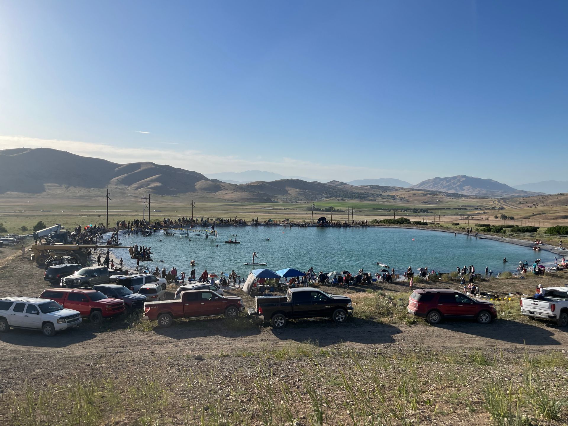 Crowded lakeside beach with swimmers, parked cars, and mountains under a clear blue sky