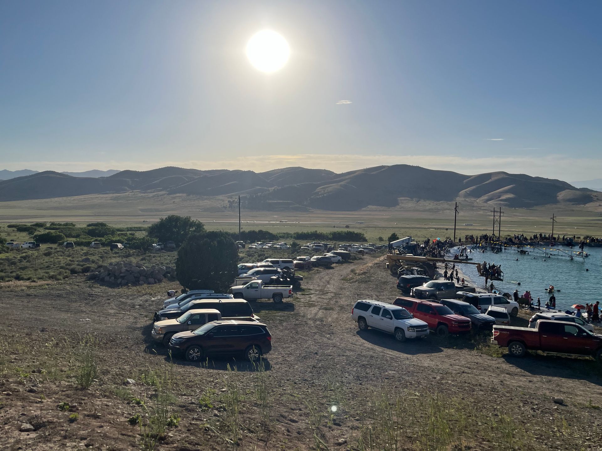 Sunlit parking lot by a lake with cars, hills, and mountains in the background