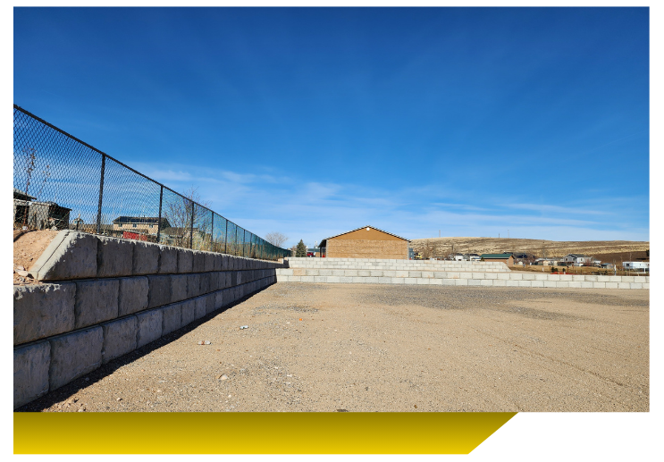 Empty gravel lot beside a stone wall and chain-link fence under a clear blue sky