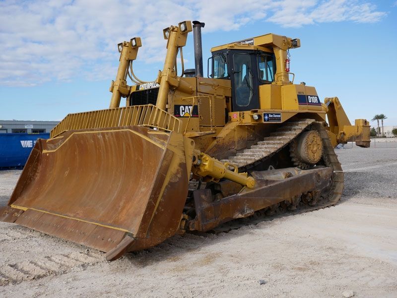 Yellow bulldozer with a large front blade parked on a gravel surface near the water under a blue sky