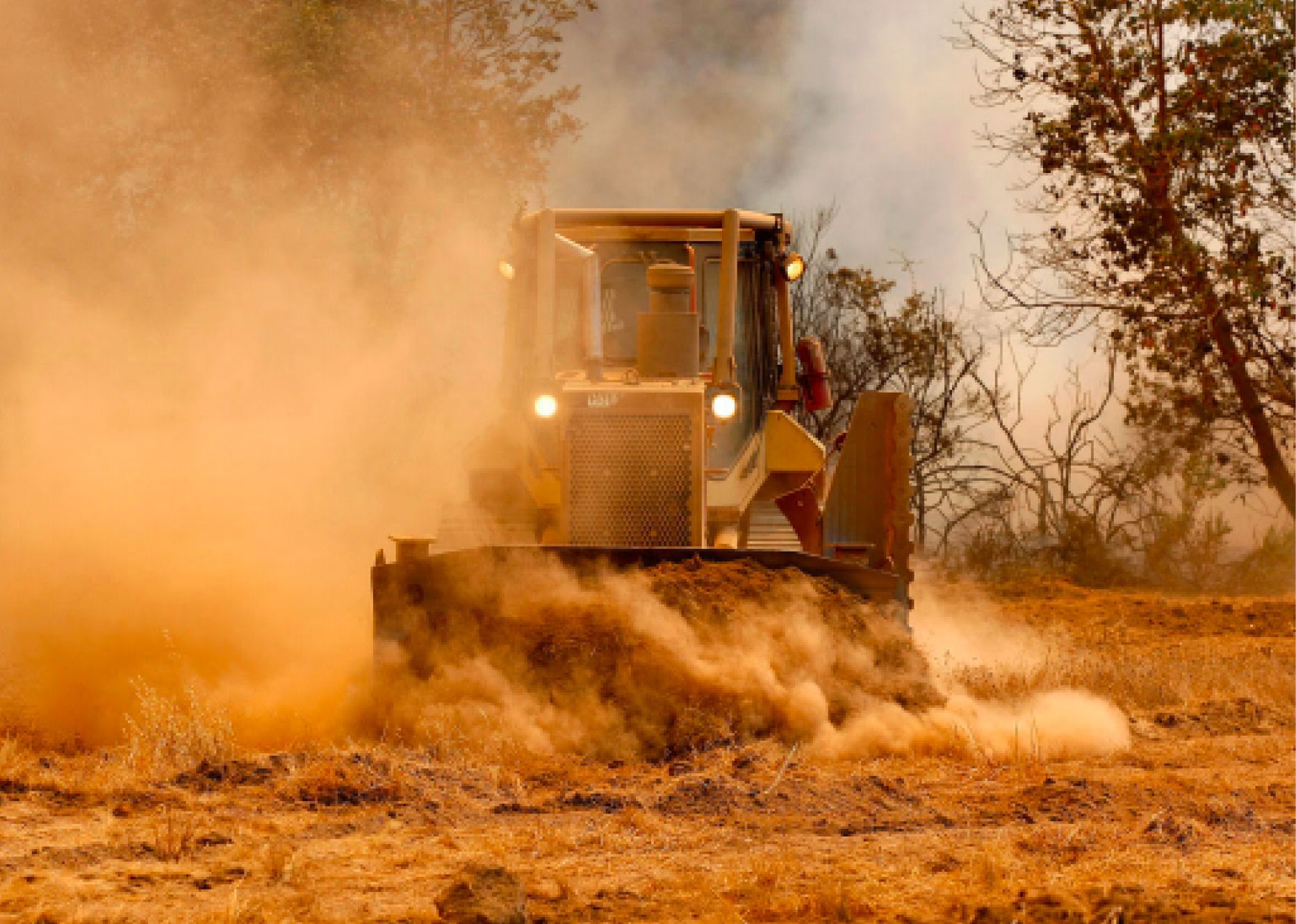 Bulldozer plowing dry field, kicking up a cloud of dust in warm sunlight