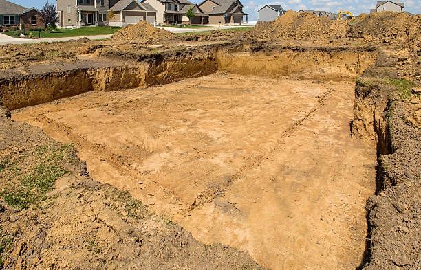 Large rectangular excavation site with exposed dirt, surrounded by mounds of soil and nearby houses in the background