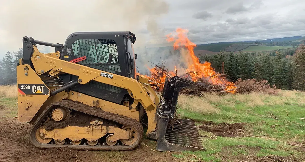 Yellow tracked bulldozer pushing burning brush in a field, flames and smoke rising beside it