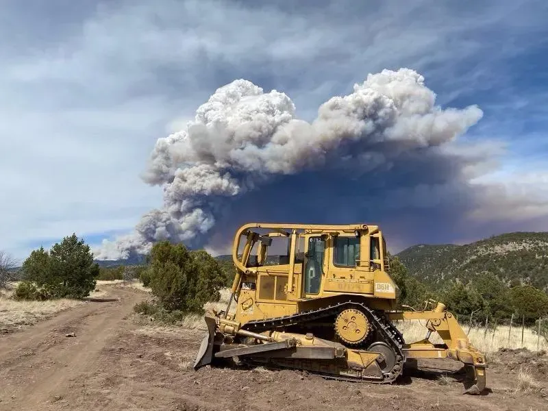 Yellow bulldozer on dirt road with a large wildfire smoke plume in the background under a blue sky