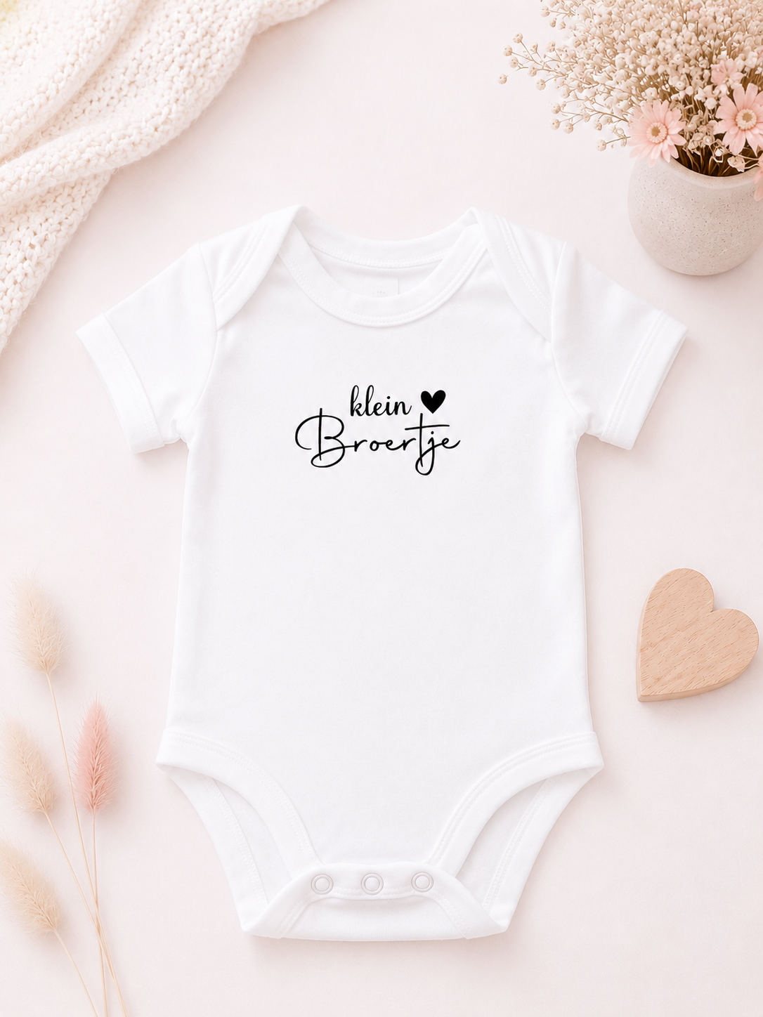Wit babyrompertje met de tekst 'Klein zusje' op een zachte wit-roze achtergrond met bloemen en houten hartje.