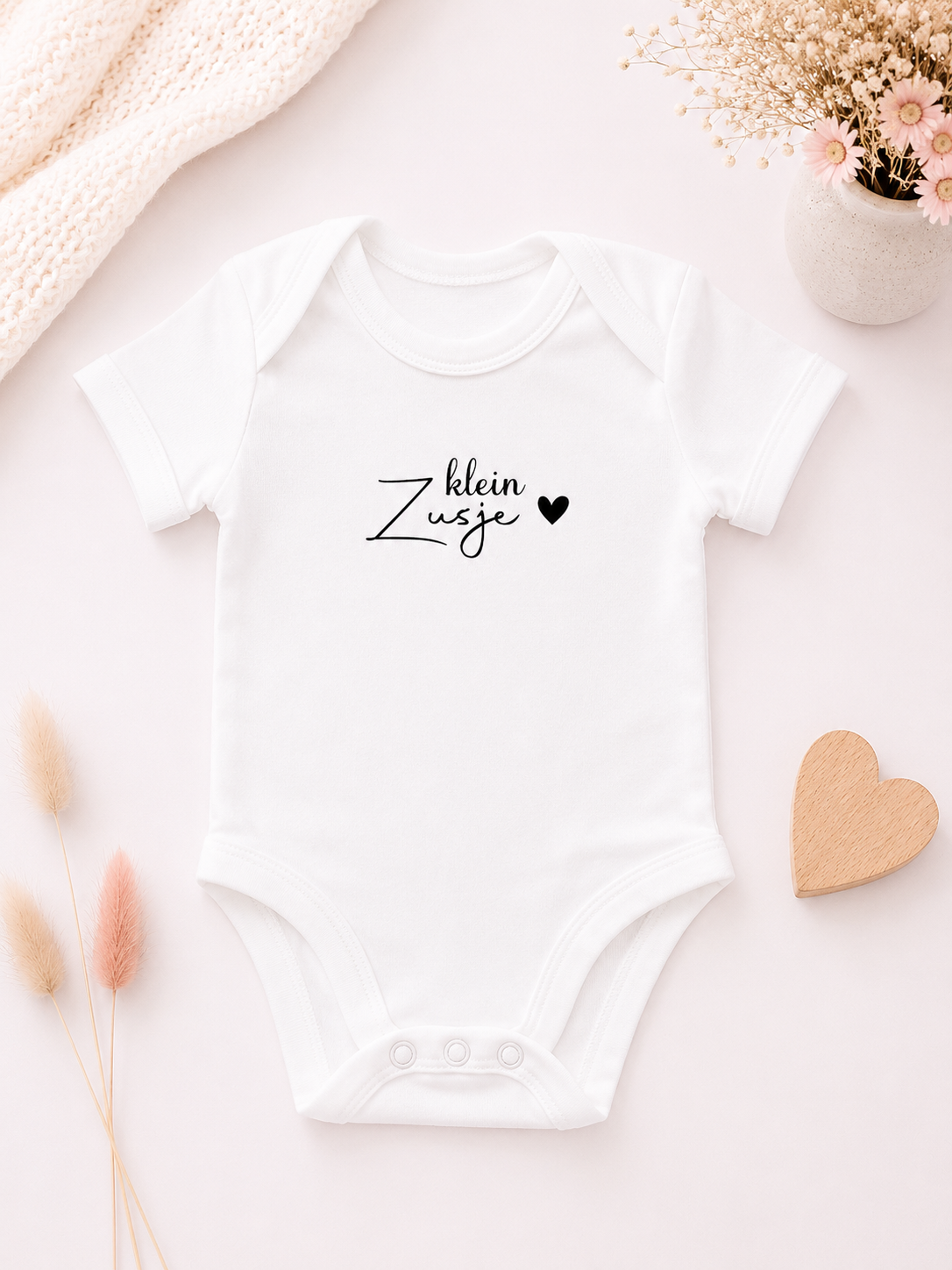 Wit babyrompertje met de tekst 'Klein zusje' op een zachte wit-roze achtergrond met bloemen en houten hartje.