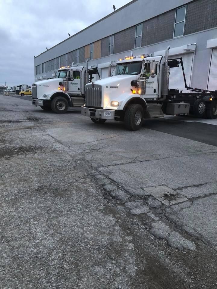 Two white dump trucks parked on cracked pavement in front of a gray industrial building. Two white dump trucks parked on cracked pavement in front of a gray industrial building.