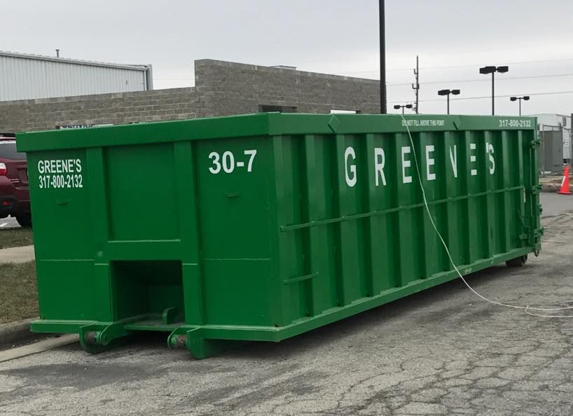 Red and green dumpsters on wheels sit side-by-side outdoors, with a backdrop of trees and a car.