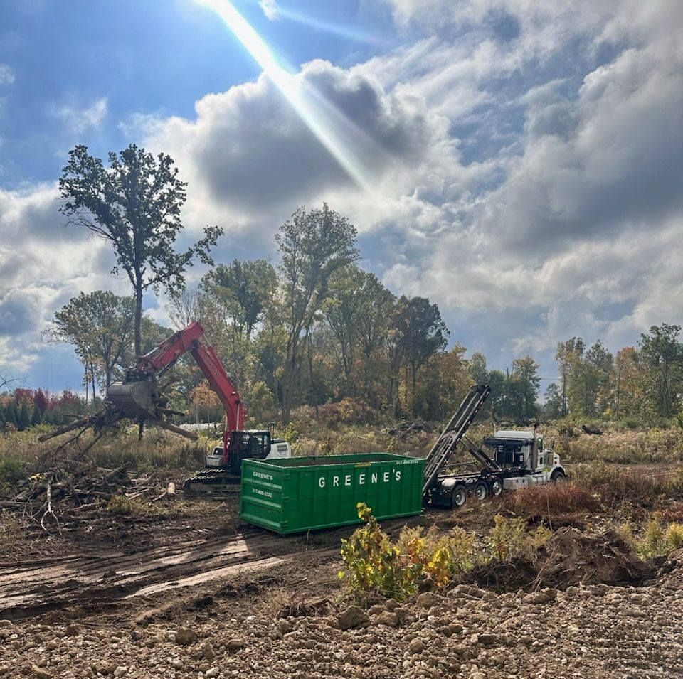 Excavator removing a tree, loading it into a green dumpster on a truck, outdoors on a cloudy day.