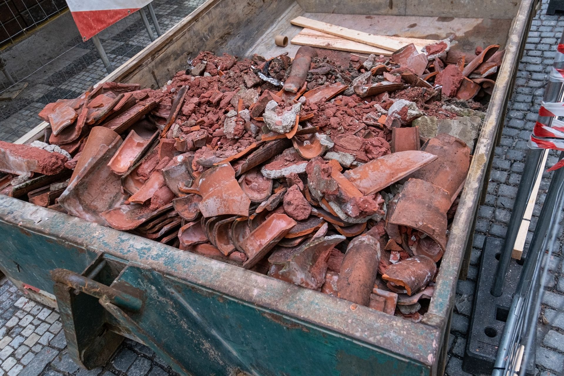 Red clay roof tiles fill a metal dumpster, likely for disposal.