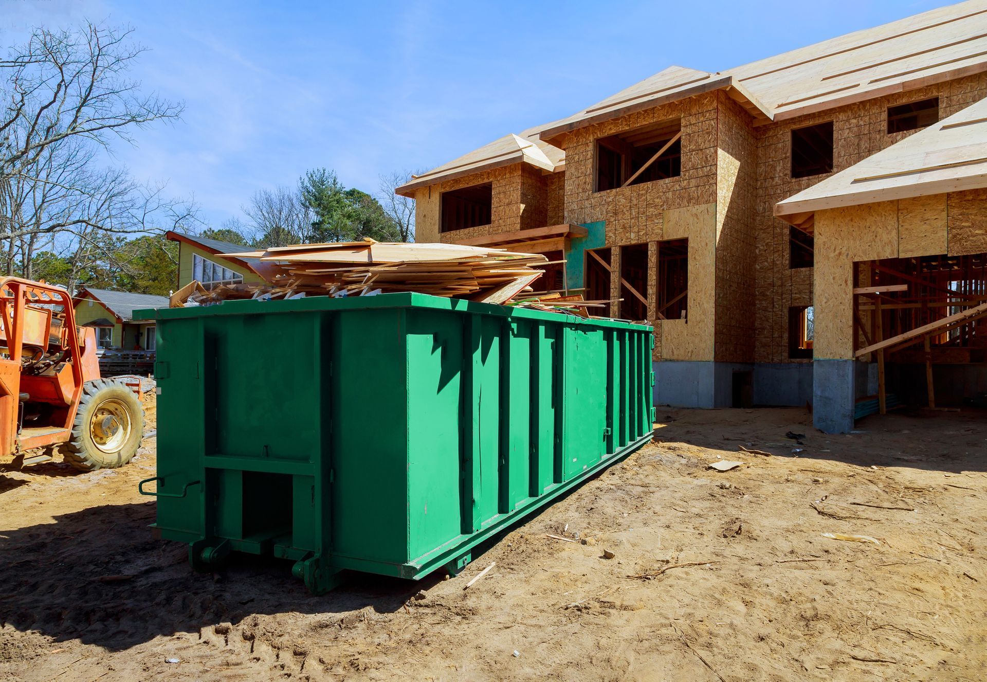 Green dumpster filled with wood debris at a construction site; unfinished house in background.