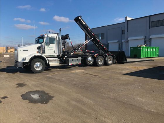 White roll-off truck lifting a dumpster in front of a gray building on a sunny day. White roll-off truck lifting a dumpster in front of a gray building on a sunny day.