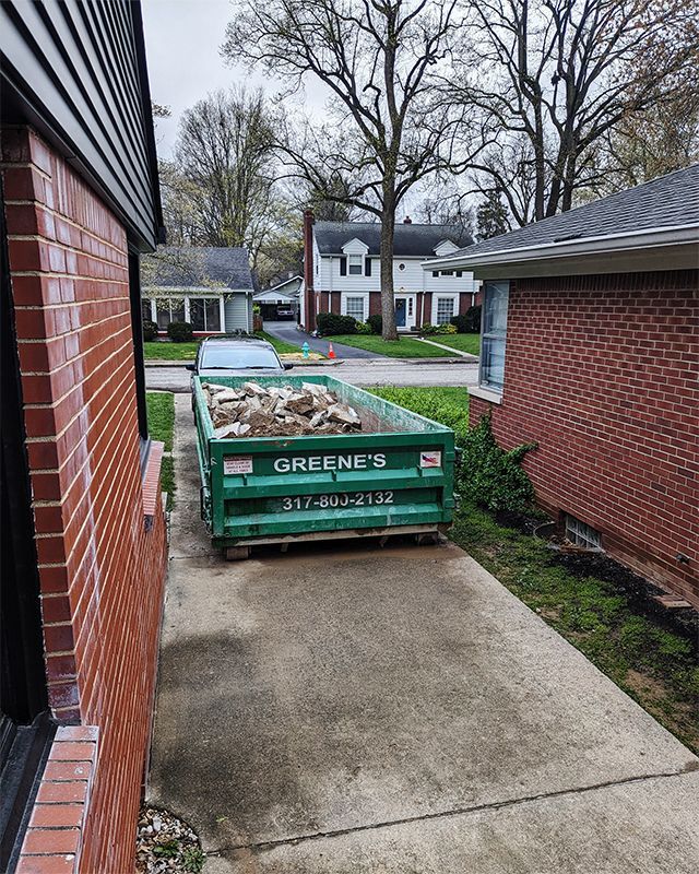 Green dumpster filled with debris in a concrete driveway between two brick buildings. Trees and houses in the background.