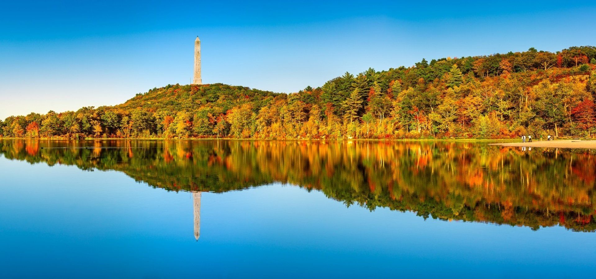 Autumn forest reflected in a calm lake, with a tall tower on the horizon.