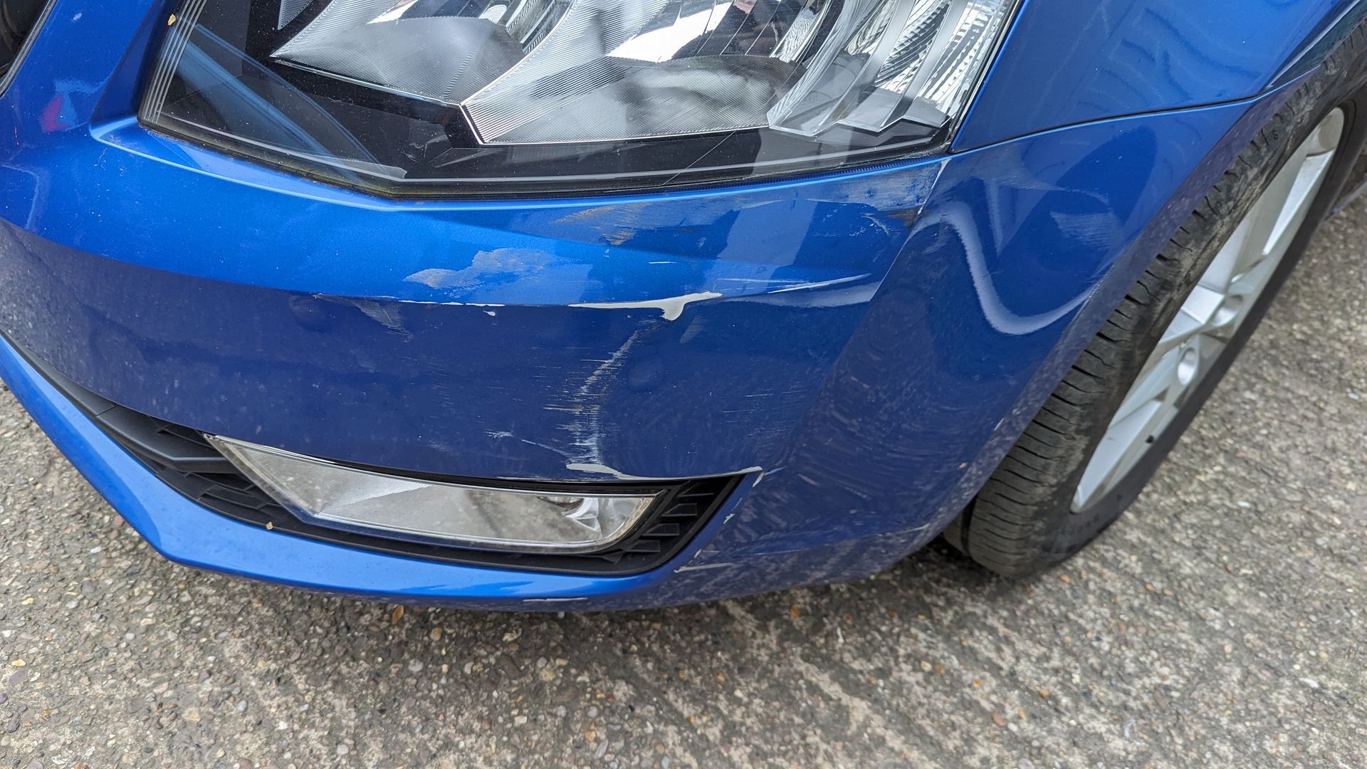 Close-up of a blue car’s front bumper and headlight with visible scrape marks and a chipped lower edge
