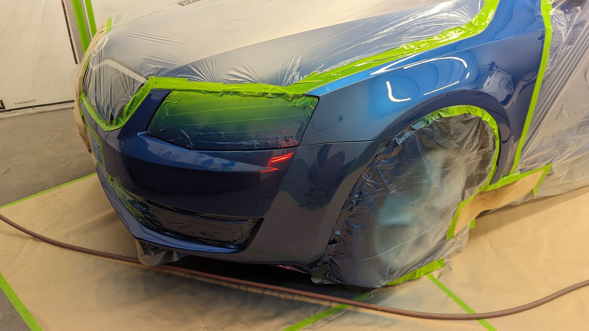 Blue sports car in a workshop, partially masked with green tape and plastic for painting or repair.