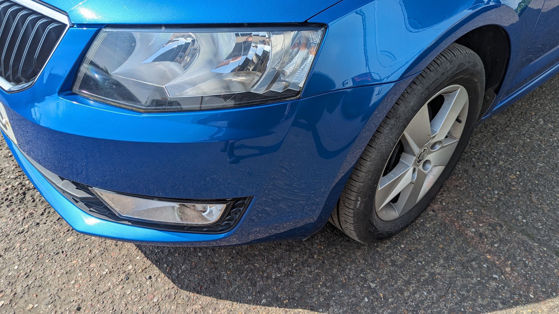 Blue car front corner with headlight, bumper, and front wheel on a road surface
