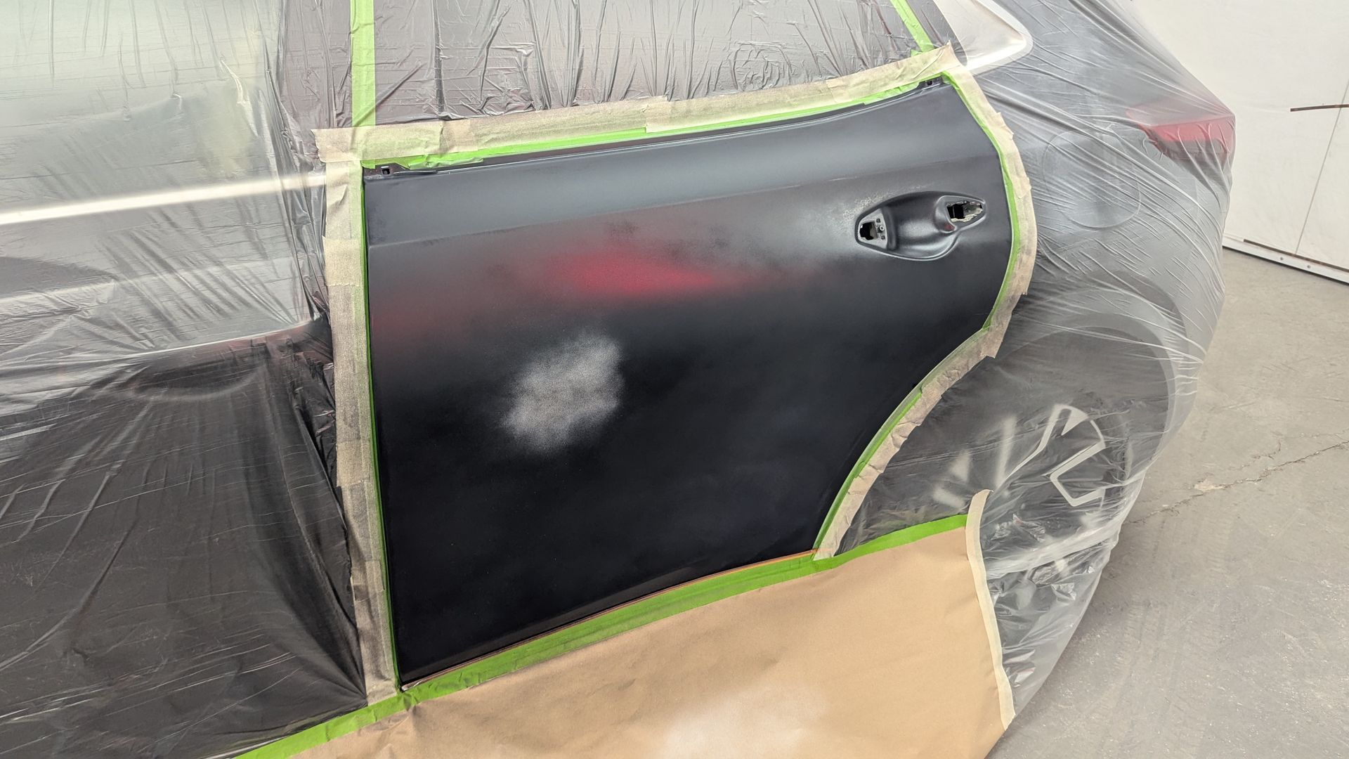 Black car door being painted, masked with tape and plastic during bodywork.