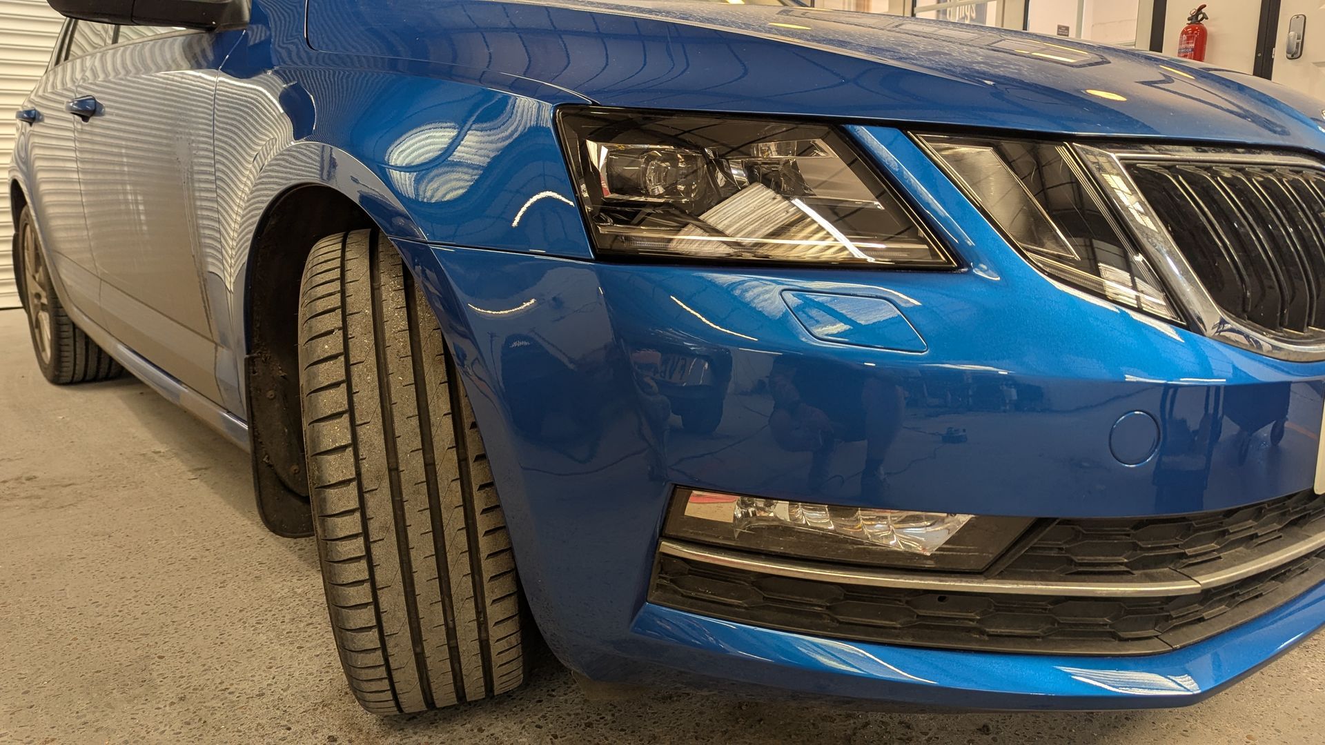 Blue car close-up in a garage, showing the front-left headlight, grille, and tire.