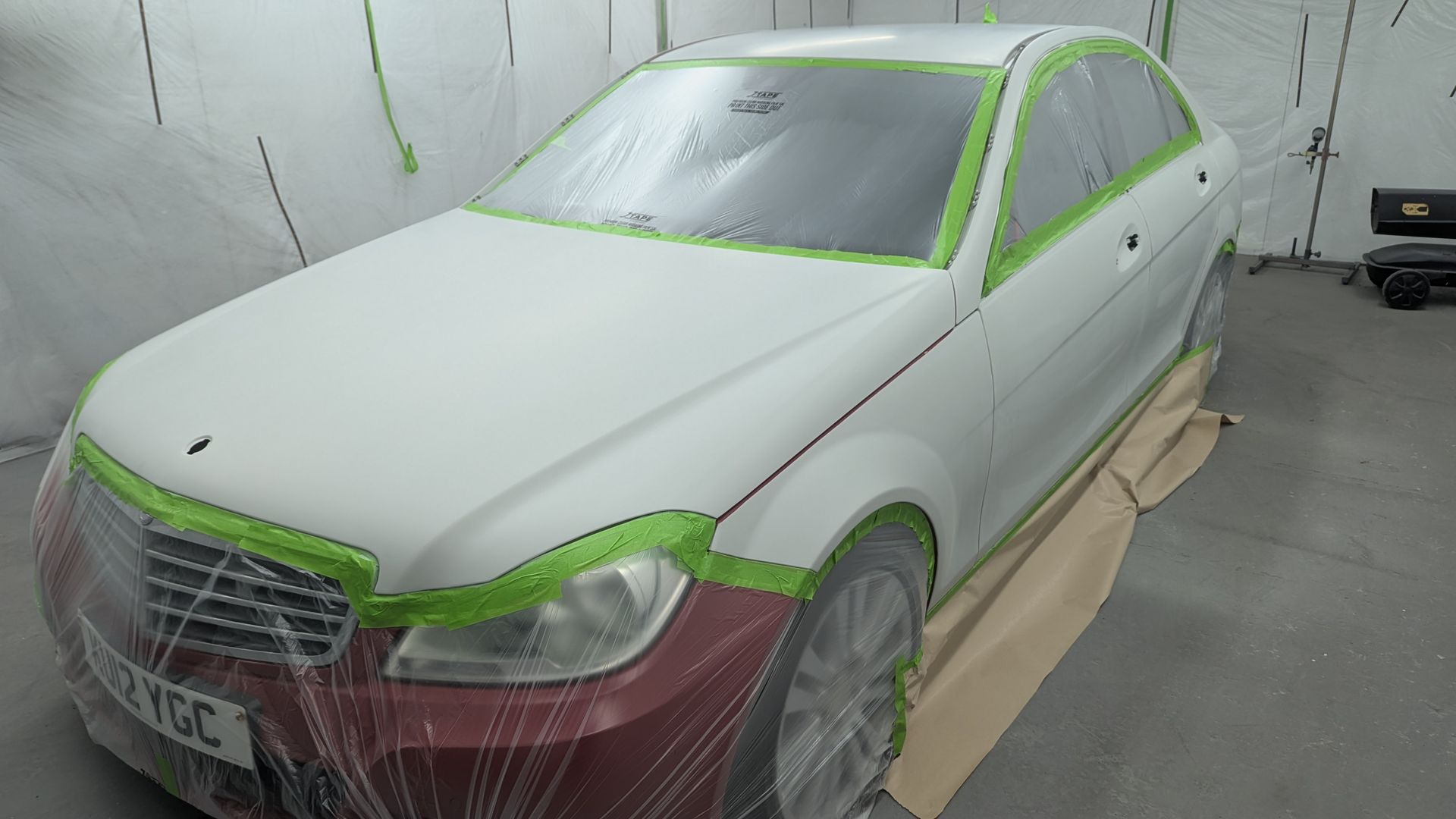 White sedan in a body shop, masked with green tape and plastic for painting repair
