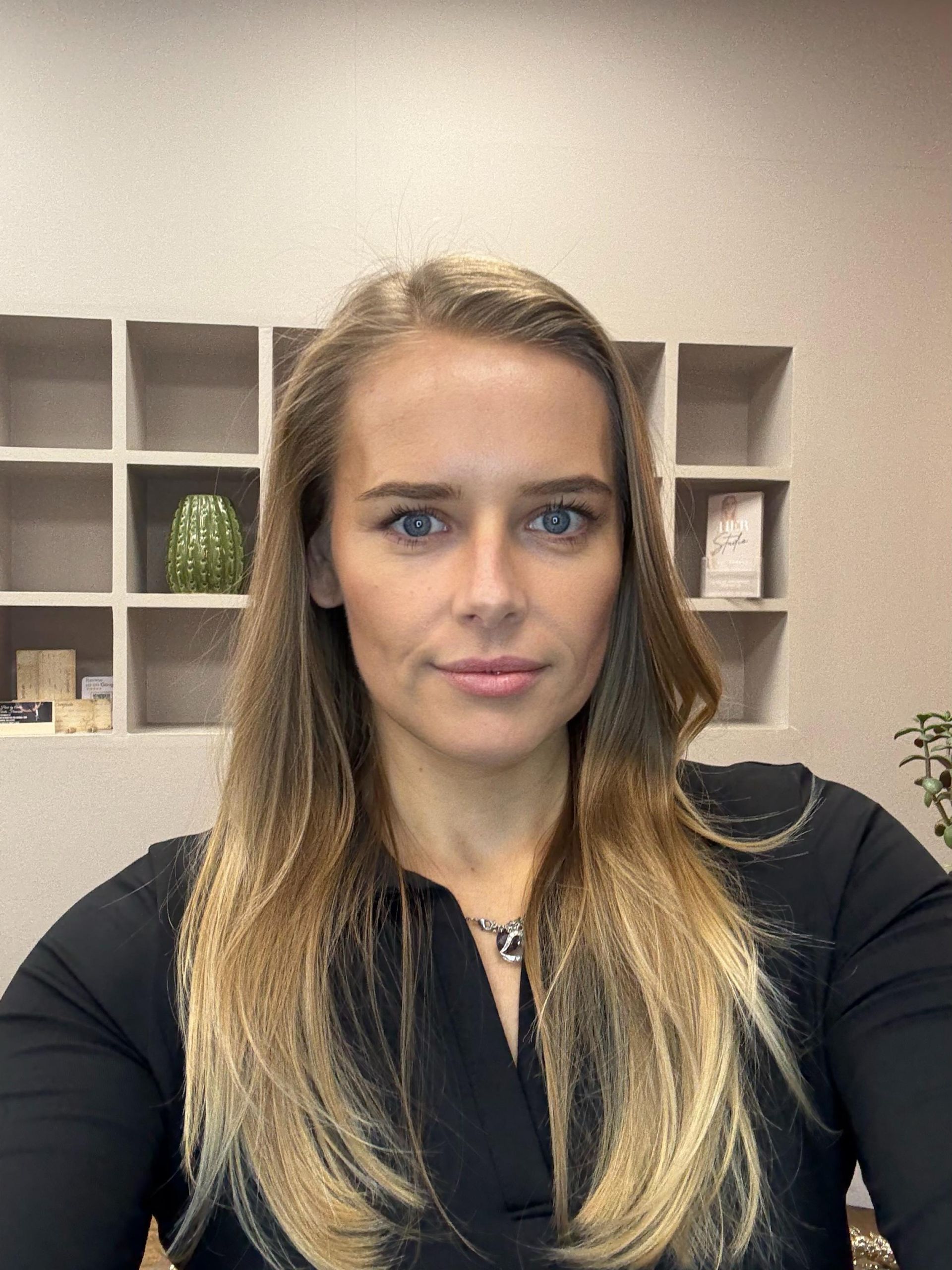 Woman with long blonde hair in a black top, seated in an office with shelves behind her.